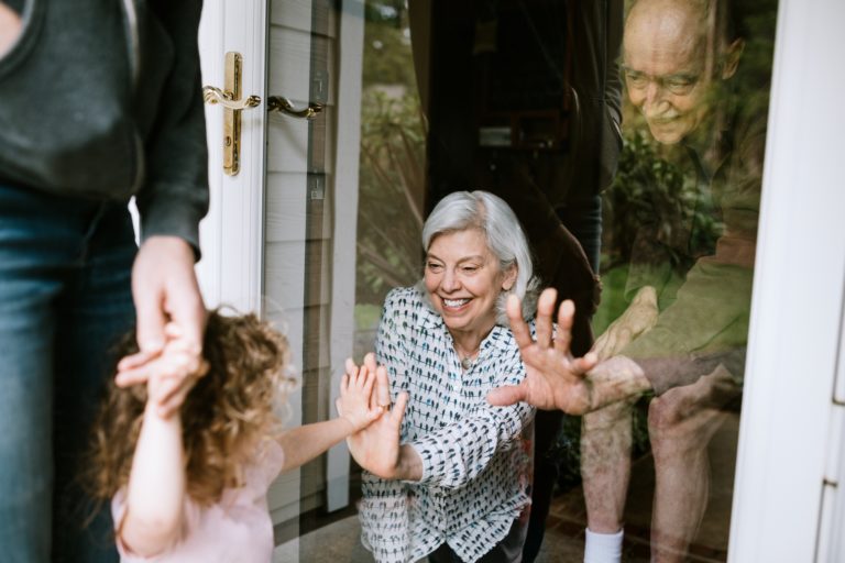 Mother with her daughter, visiting senior parents but observing social distancing with a glass door between them. The granddaughter puts her hand up to the glass, the grandfather and grandmother doing the same